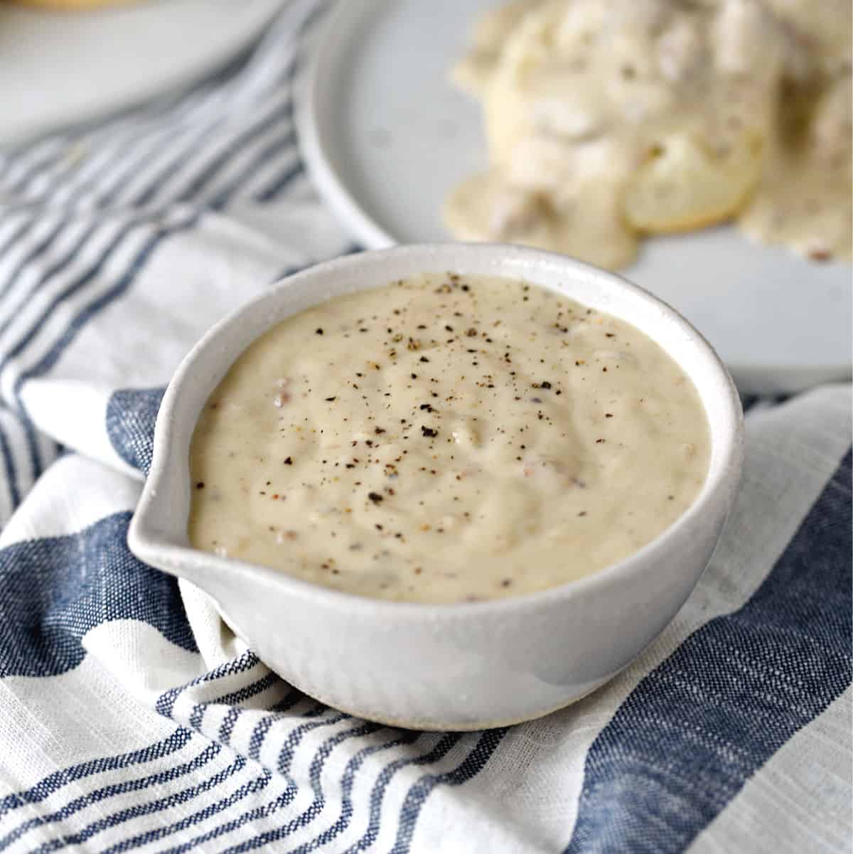 a white bowl filled with thick and creamy sausage gravy with a plate of biscuits topped with gravy in the background.