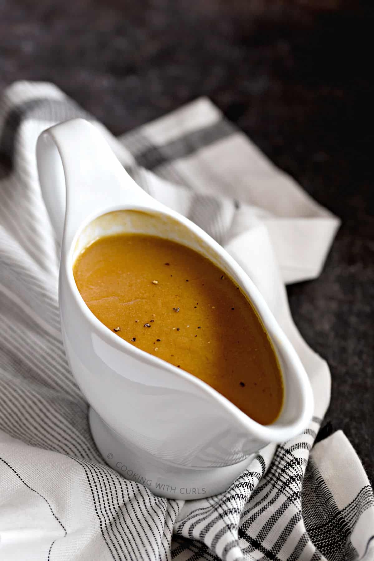 Rich, brown turkey gravy sprinkled with black pepper in a white gravy boat sitting on a black and white striped napkin.