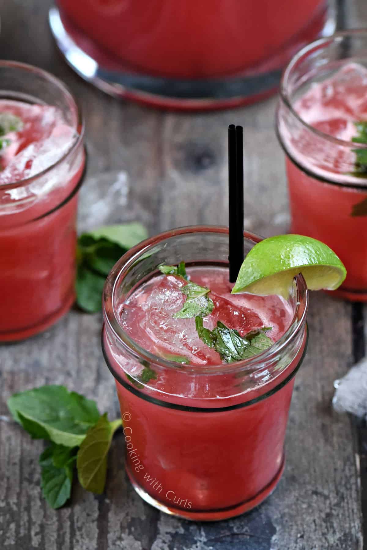 Three watermelon mojitos with lime wedges and mint leaves with a pitcher in the background.