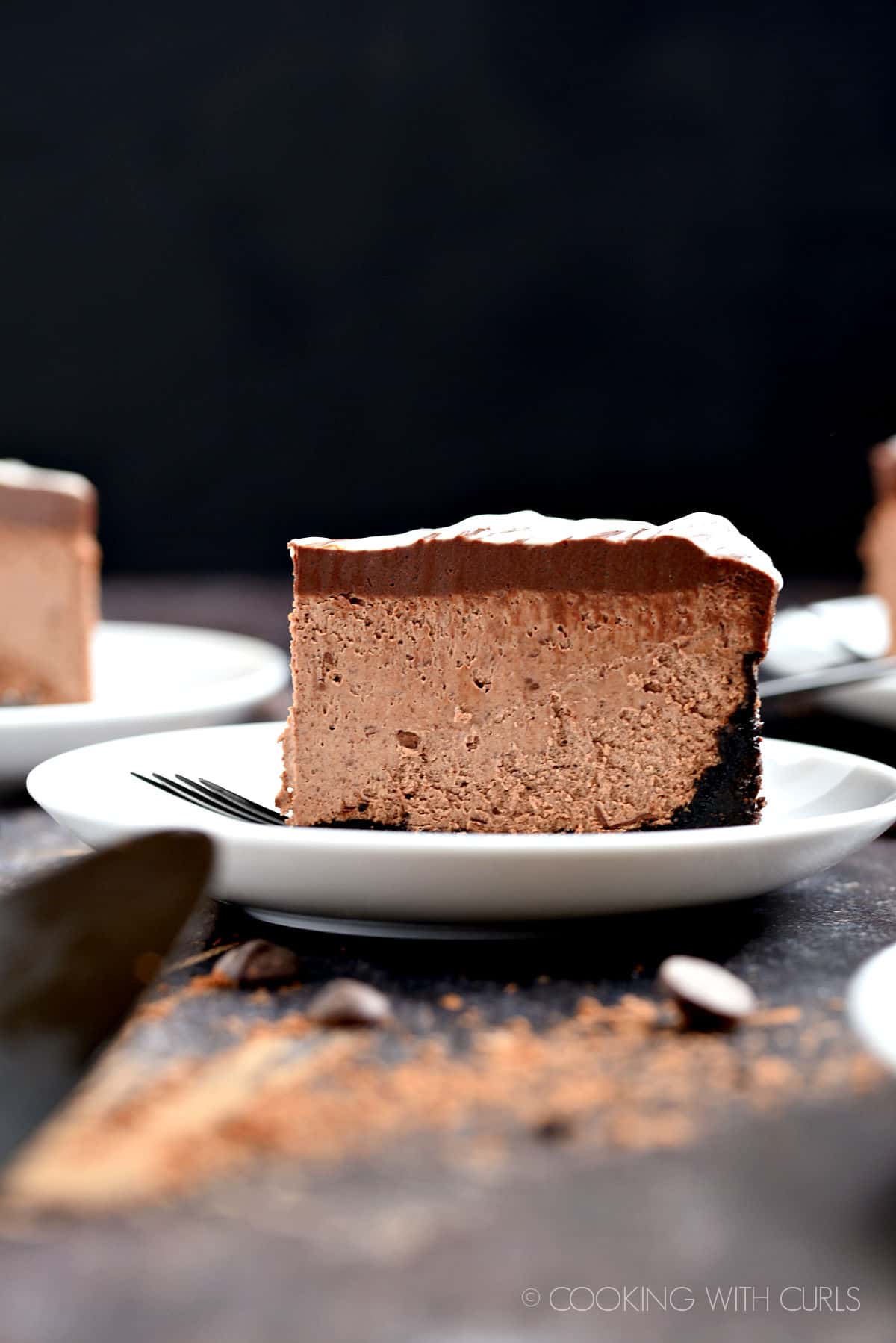 A slice of chocolate cheesecake on a small white plate with a black fork.