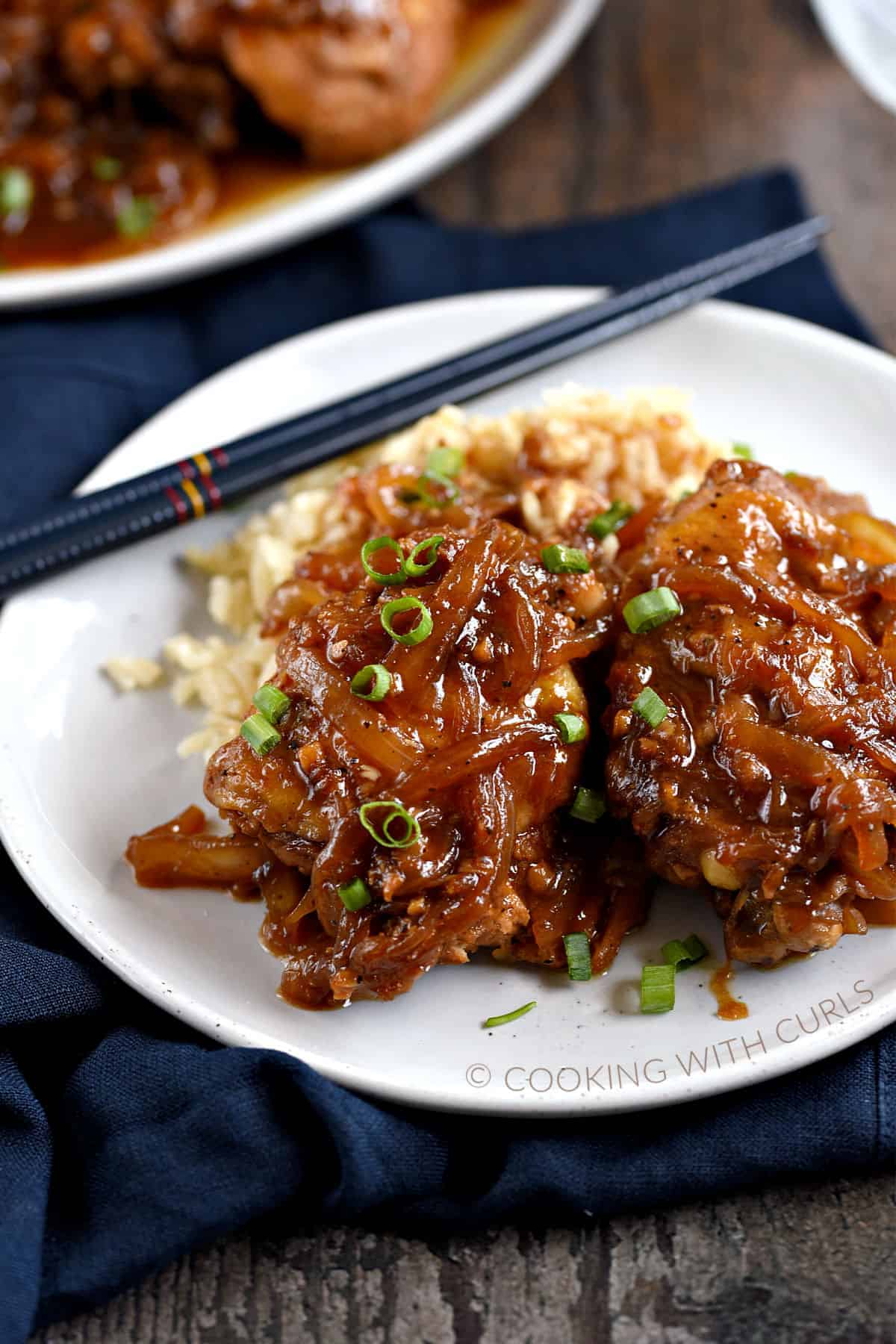 Two chicken thighs covered with a brown onion sauce and diced green onions on a bed of rice with chopsticks in the upper left corner of the plate.