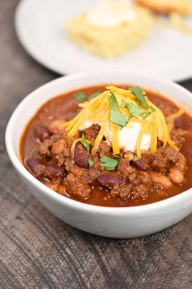 Beef Chili in a white bowl topped with sour cream, shredded cheddar cheese, and cilantro leaves with a plate of cornbread in the background.