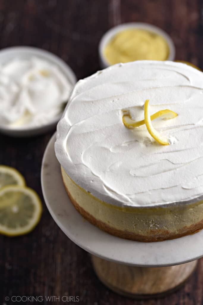 Instant Pot Lemon Cheesecake topped with lemon curd and whipped cream sitting on a marble topped cake plate with bowls of whipped cream and lemon curd in the background.