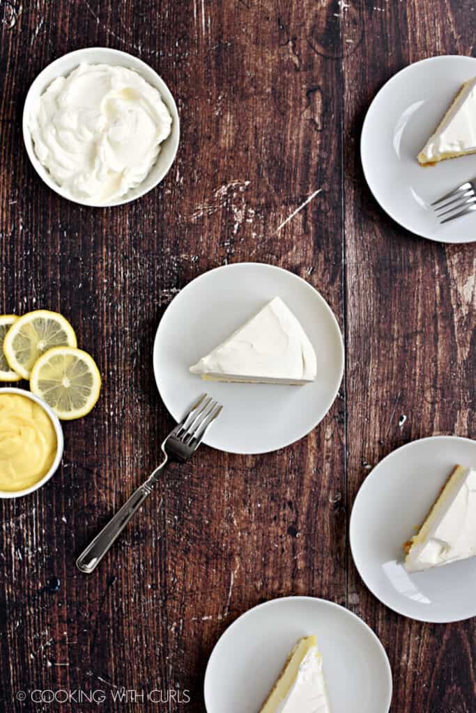 Looking down on four slices of cheesecake on small white plates, with bowls of whipped cream and lemon curd in the background.