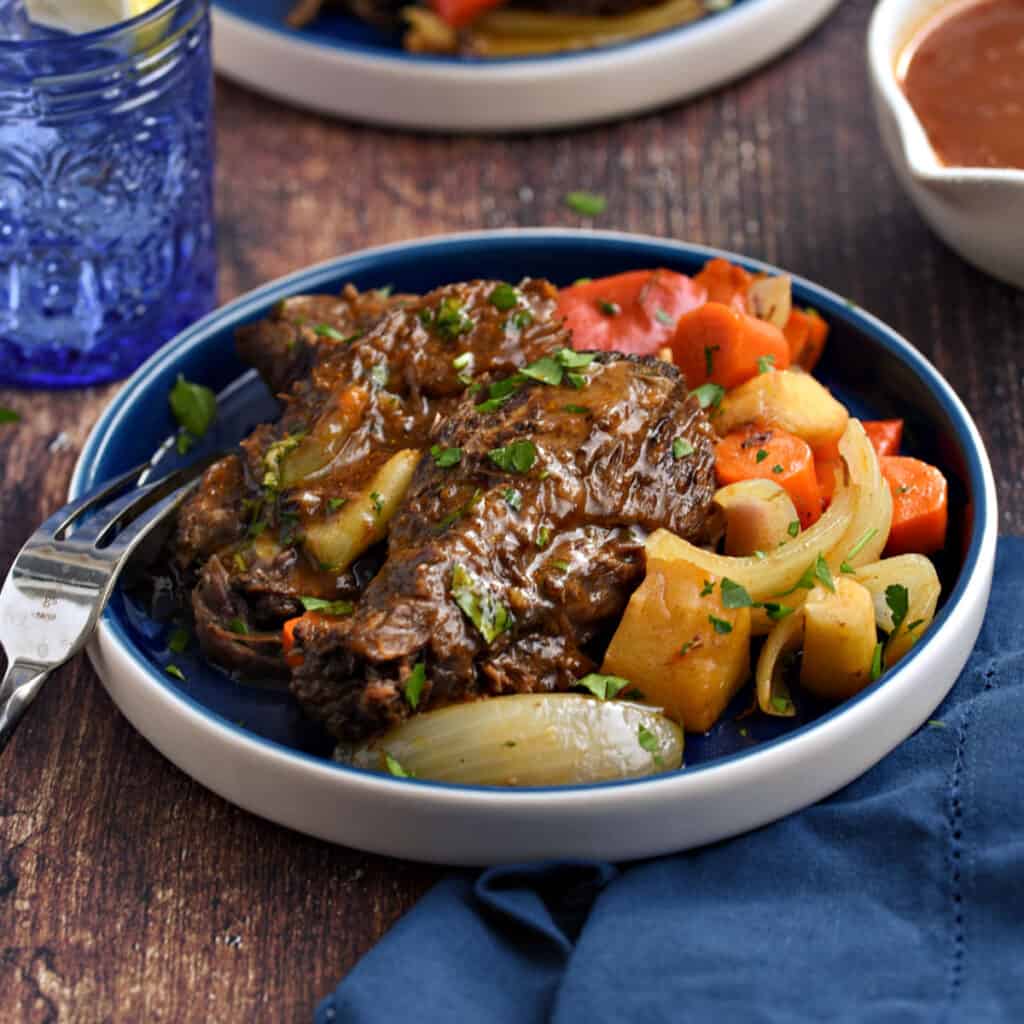 Pot roast with carrots, onions and parsnips on a blue plate with a blue glass and blue napkin in the background.