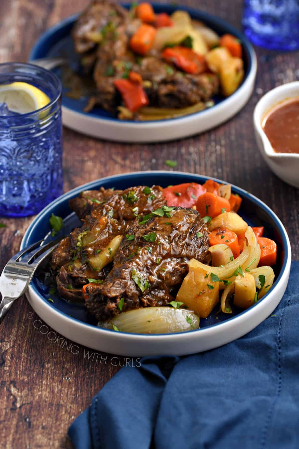 Pot roast topped with gravy and served with chopped red bell peppers, carrots, parsnips and onion wedges on a blue plate.