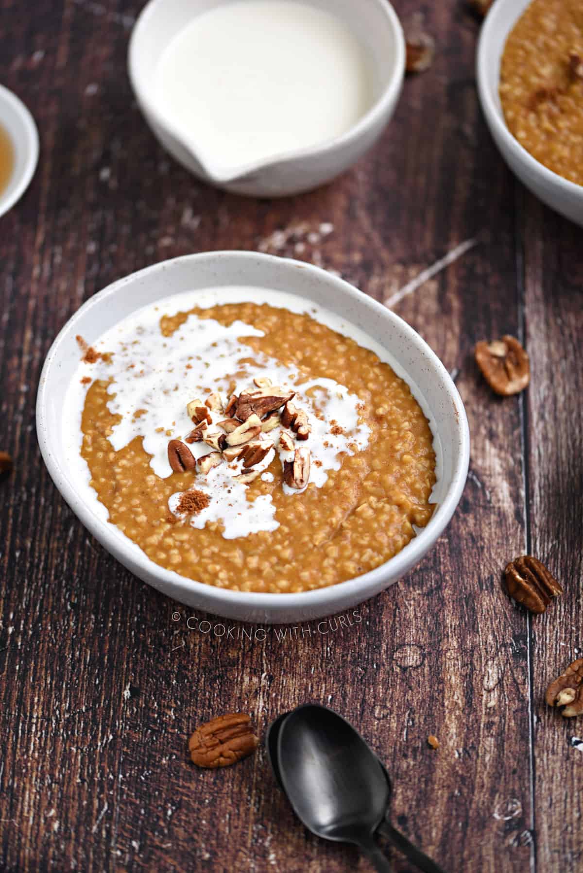 Pumpkin oatmeal in a bowl topped with pecan pieces and cream, with a bowl of cream and second bowl of oatmeal in the background.