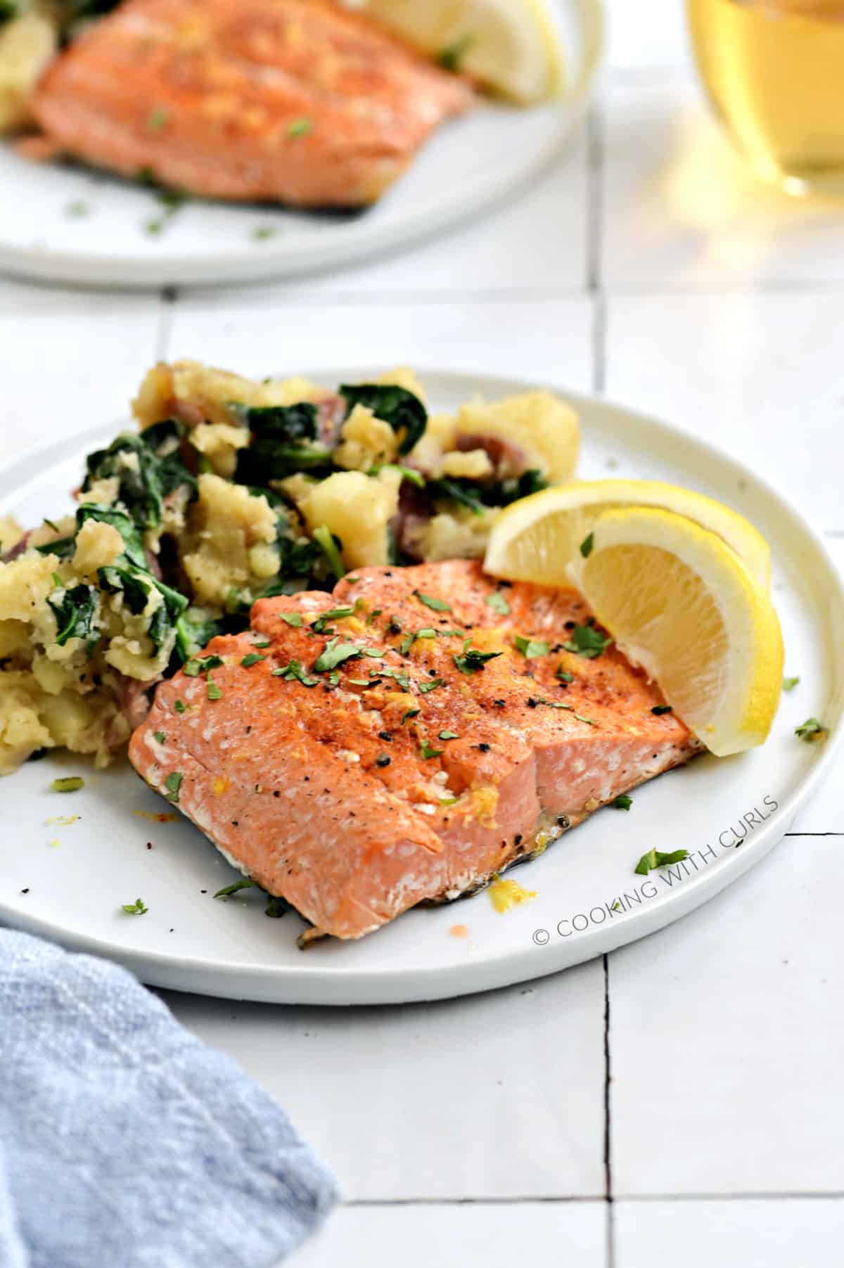 A salmon filet and mashed potatoes mixed with baby spinach on a white plate with two lemon wedges.