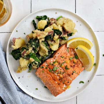 A salmon filet and mashed potatoes mixed with spinach on a white plate with two lemon wedges.