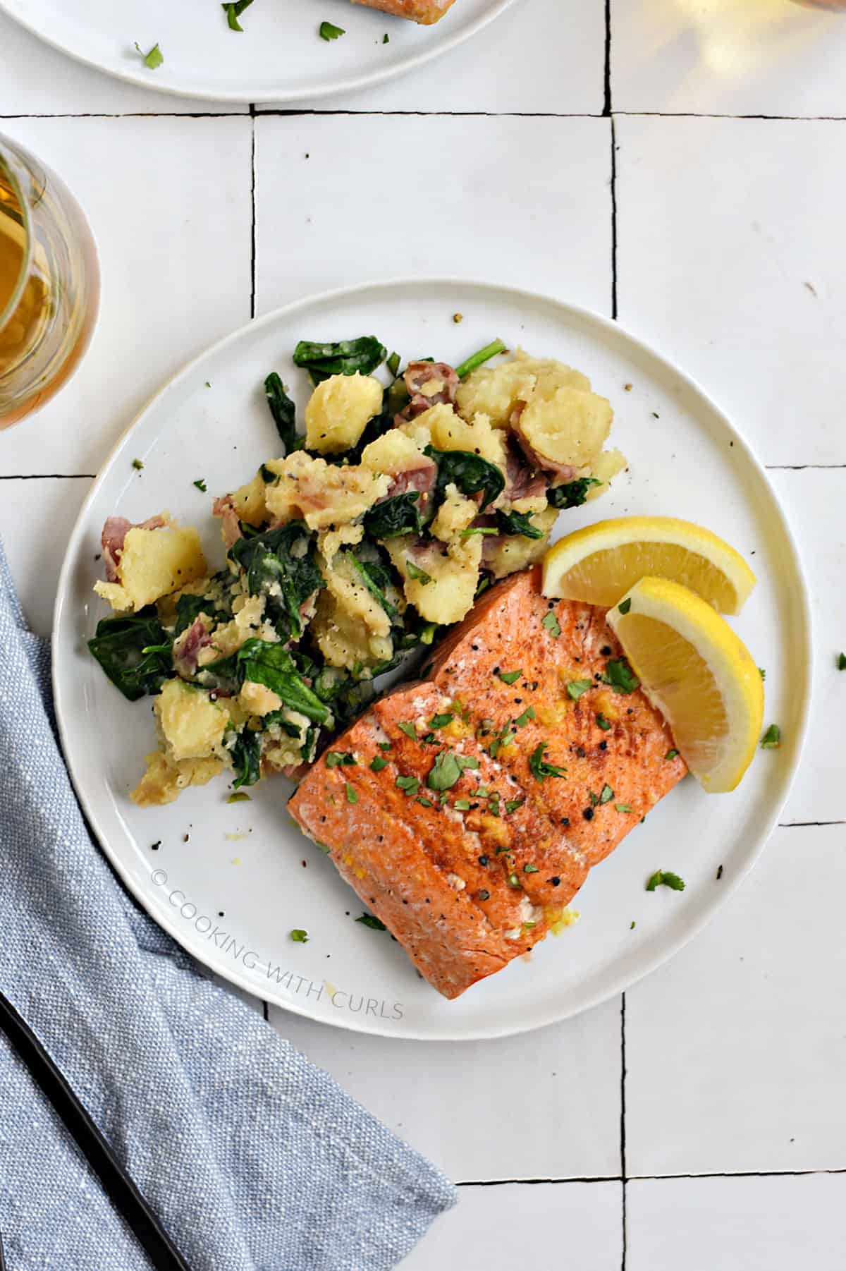 Looking down on a salmon filet and mashed potatoes mixed with baby spinach on a white plate with two lemon wedges.