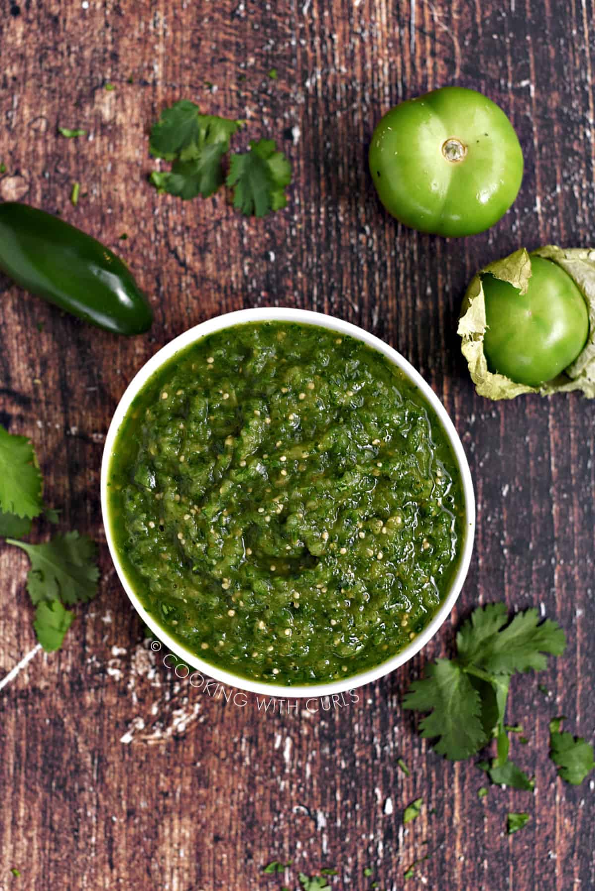 Looking down on a white bowl filled with green salsa verde surrounded by tomatillos, jalapeno pepper and cilantro leaves.