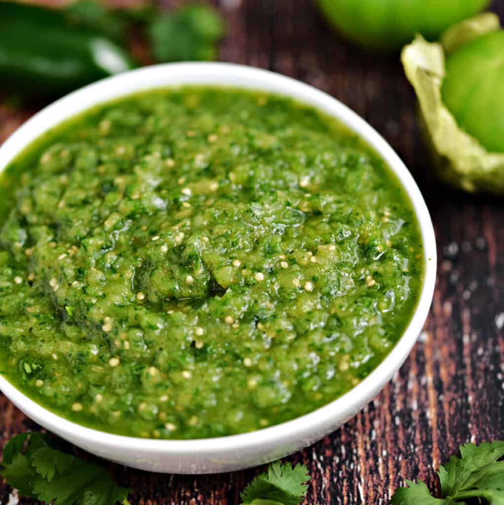 Blended, Instant Pot Salsa Verde in a white bowl surrounded by cilantro, tomatillos and a jalapeno pepper.