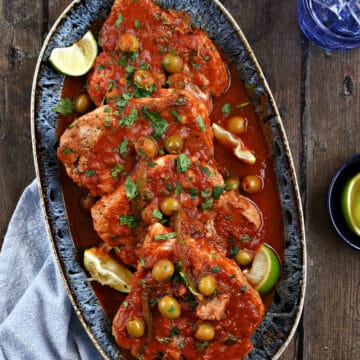 Looking down on four pork chops on a platter topped with tomato, caper, pepper, and olive sauce.