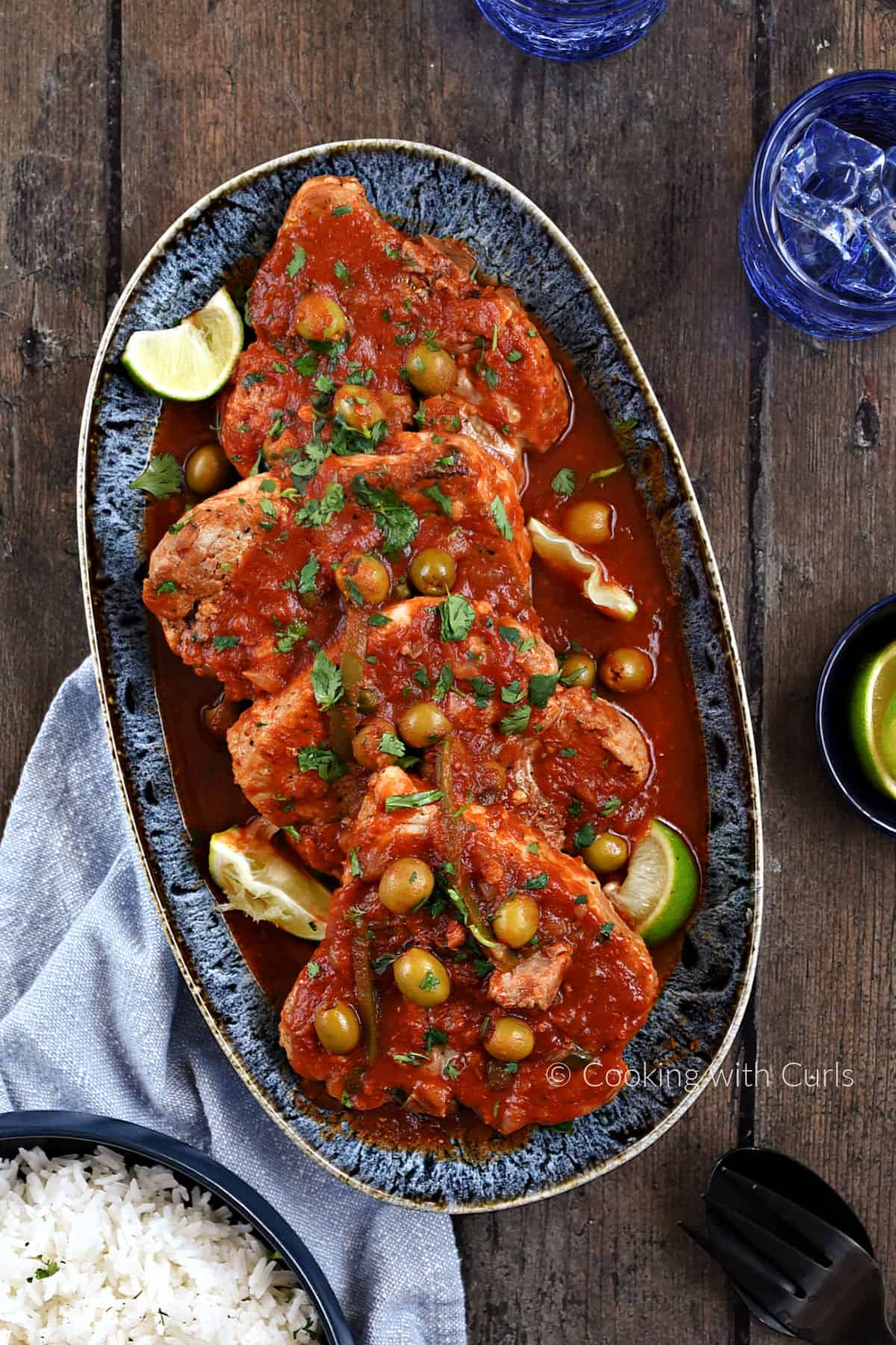 Looking down on four pork chops on a platter topped with tomato, caper, pepper, and olive sauce.