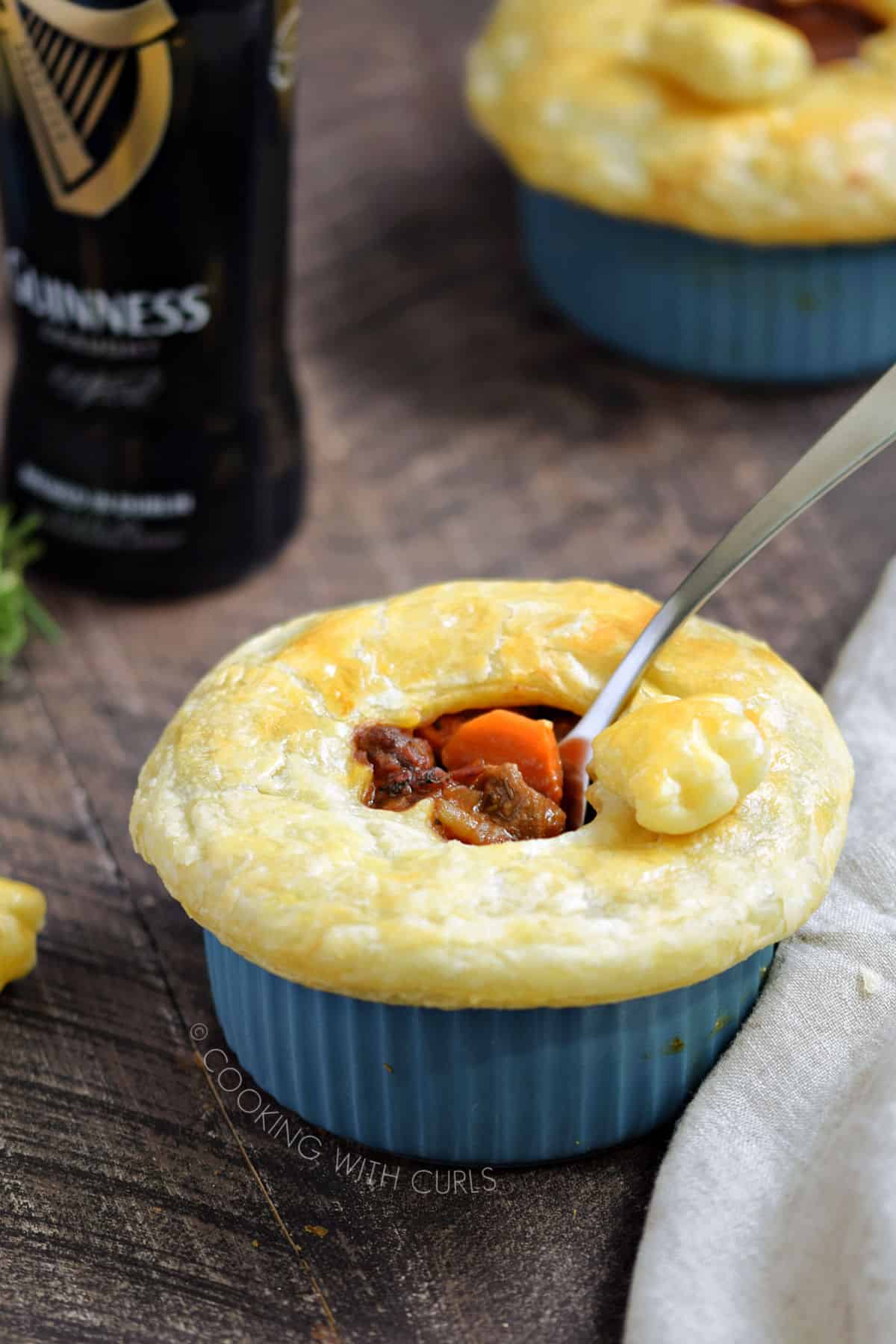 Puff pastry topped beef stew in a blue ramekin with a bottle of Guinness and a beige napkin in the background.