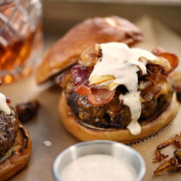 A burger topped with grilled onions, bacon, and whiskey mayonnaise on a pretzel bun with a glass of whiskey in the background.