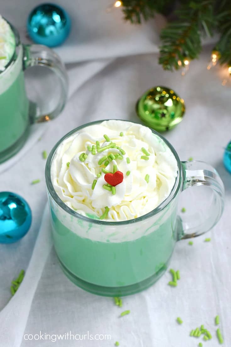 Two glass mugs filled with green hot chocolate topped with whipped cream, green sprinkles, and a red heart.