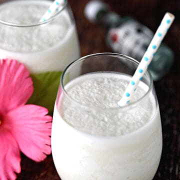 two glasses filled with creamy pina coladas and a pink hibiscus flower between them.