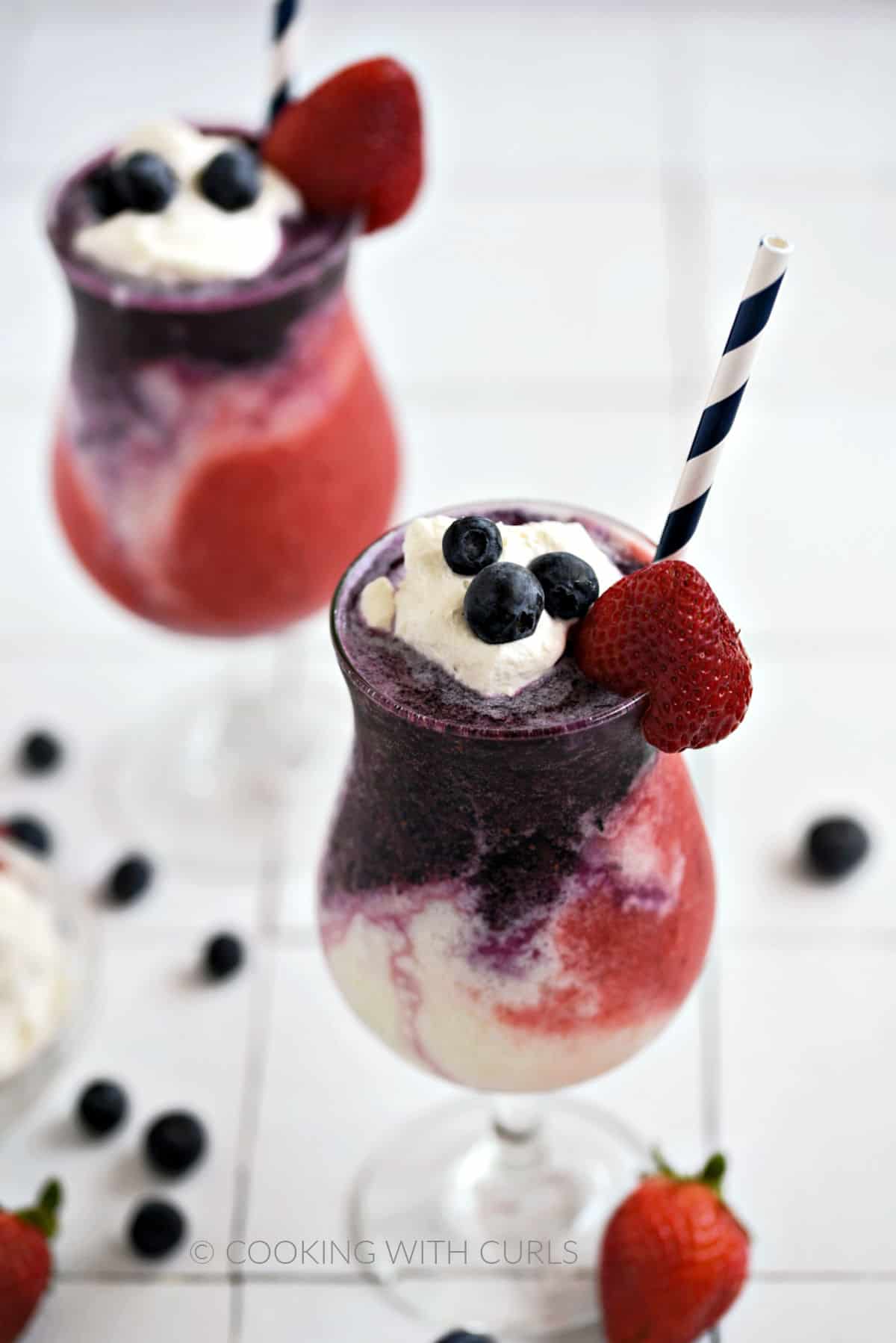 Blueberry, white, and strawberry layers topped with whipped cream, fresh blueberries and a strawberry in two hurricane glasses with a blue and white striped straw and scattered berries around the base.