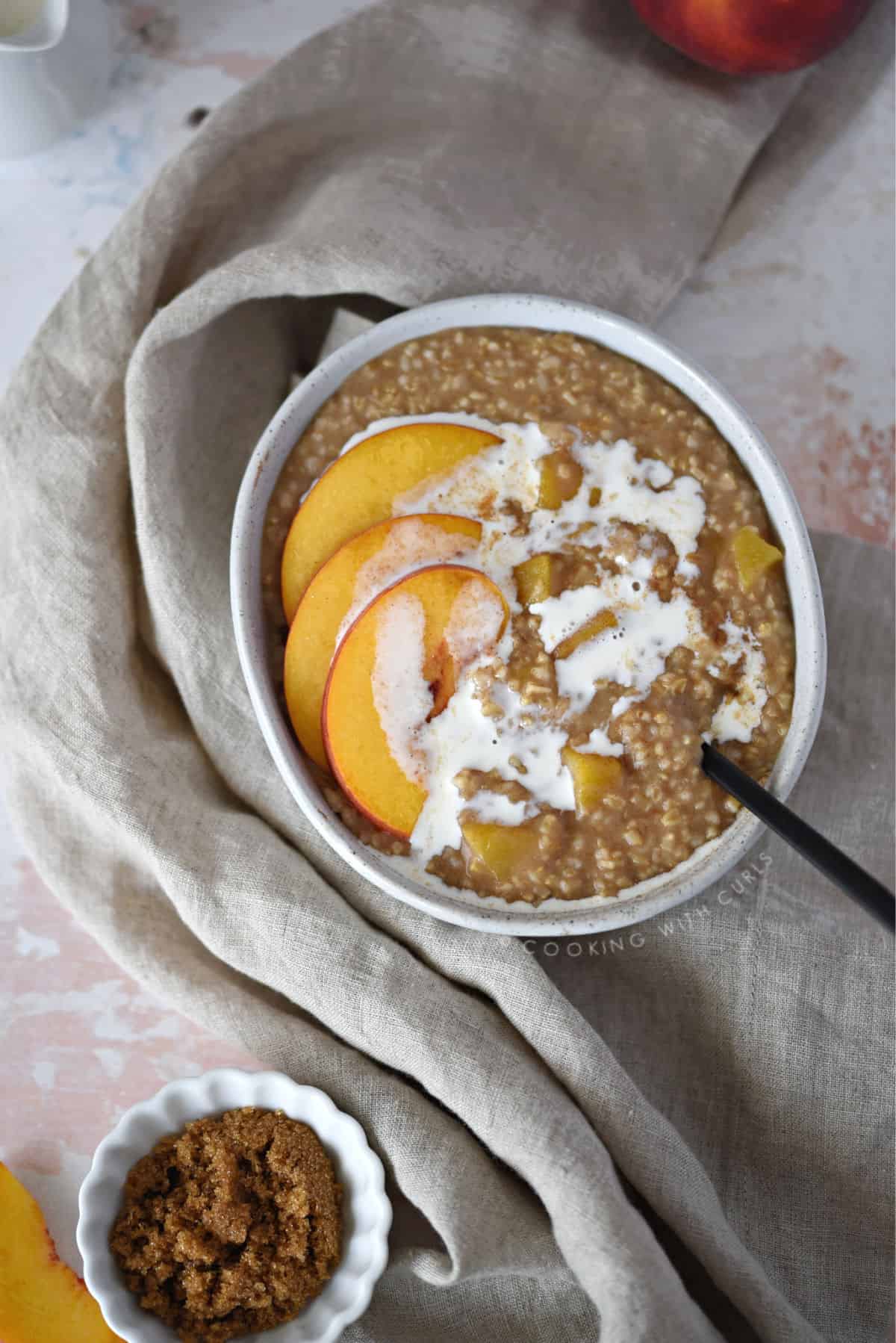 looking down on a bowl of steel cut oats topped with peach slices and cream, with a small bowl of brown sugar in the bottom left corner and the edge of a full peach in the upper right corner.