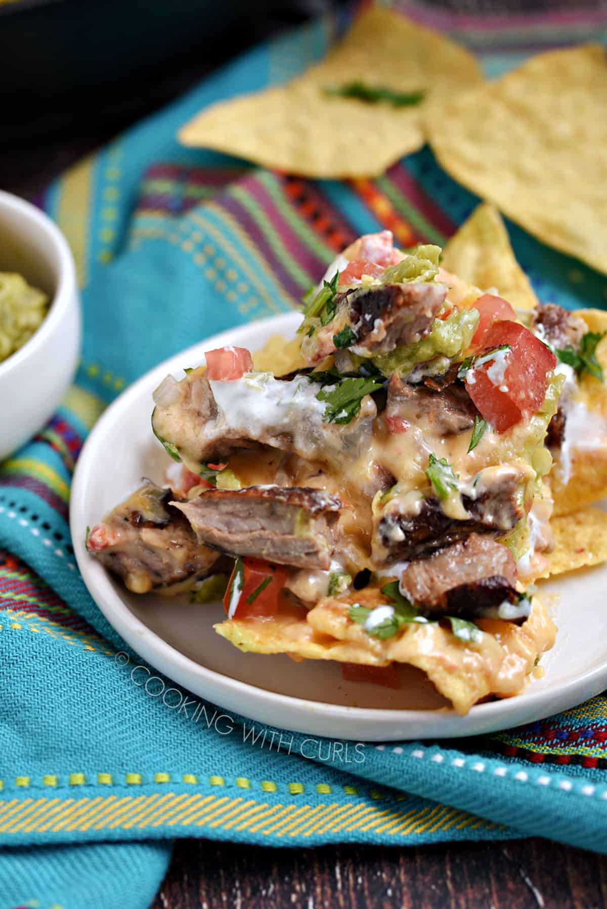 Tortilla chips topped with melted cheese, queso, guacamole, diced tomatoes, cilantro and onions on a small white plate.