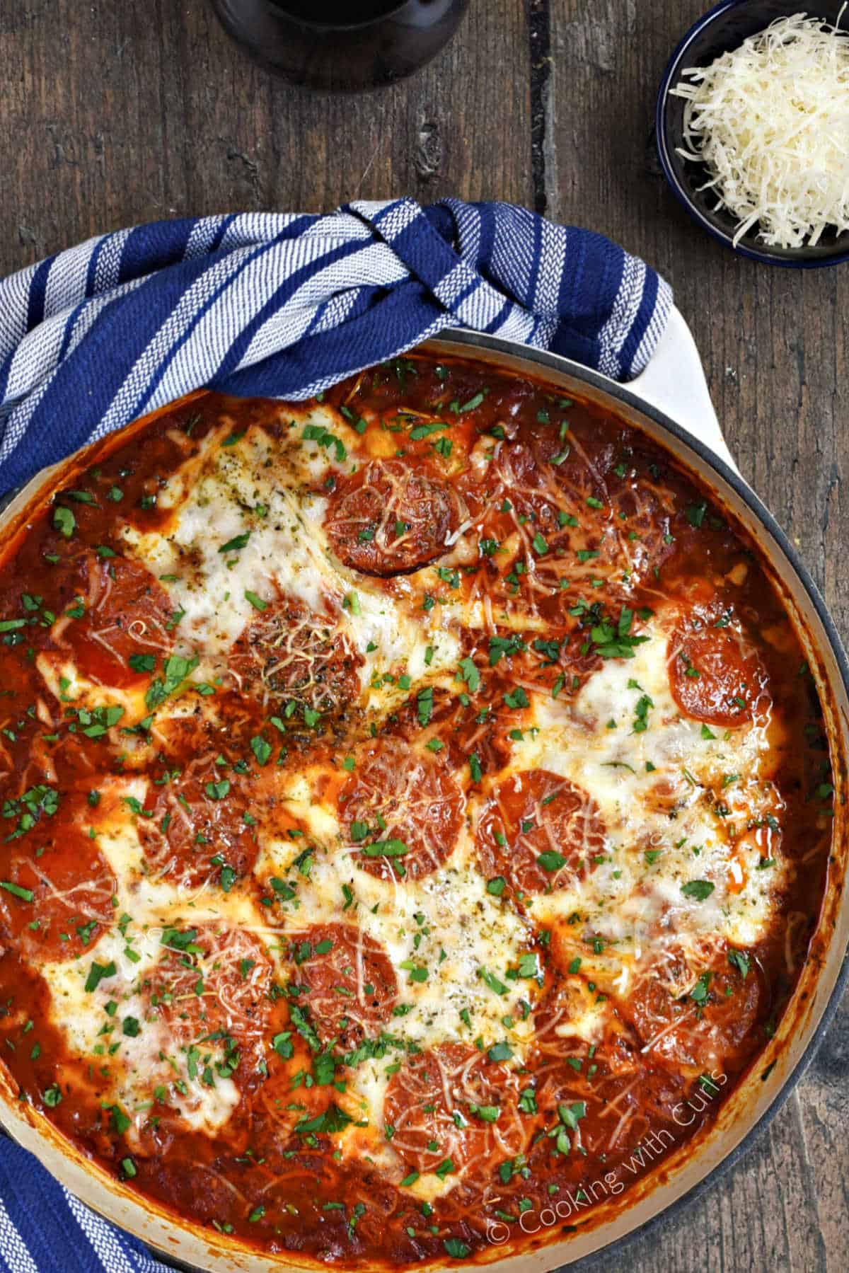 Looking down on a pizza chicken bake in a large skillet with a bowl of grated Parmesan in the upper right corner.