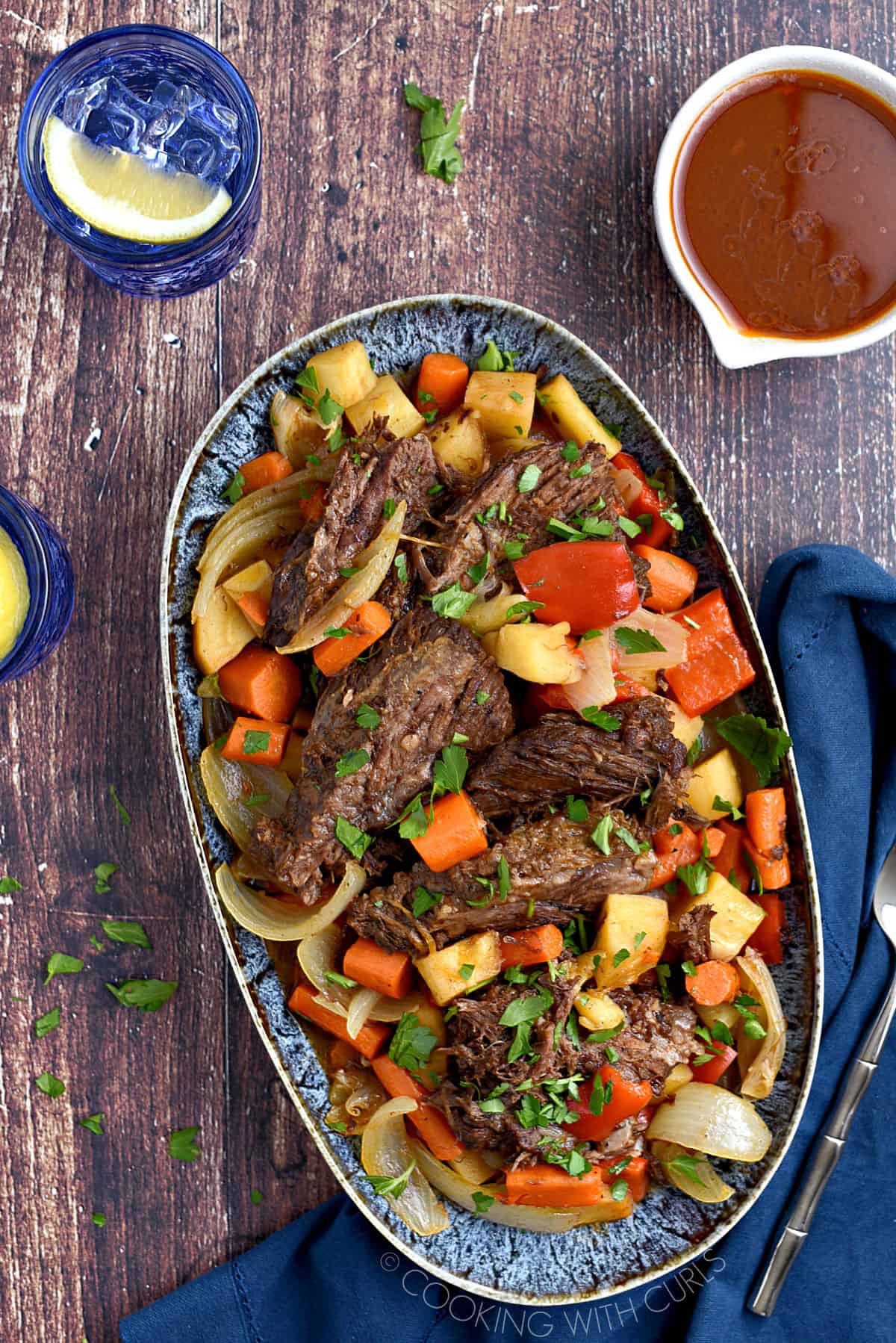 Looking down on a platter filled with Instant Pot Moroccan Pot Roast and vegetables with a blue glass of water and a white bowl of gravy in the background.