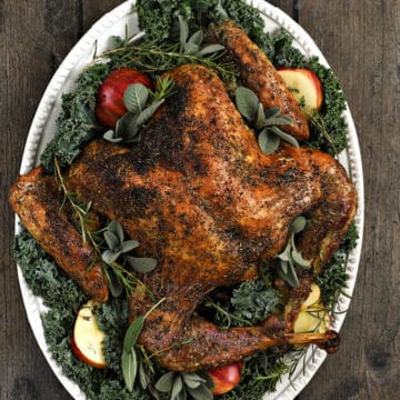 Looking down on a spatchcock turkey recipe on an oval platter covered in kale, sliced apples, sage, rosemary, and thyme sprigs.