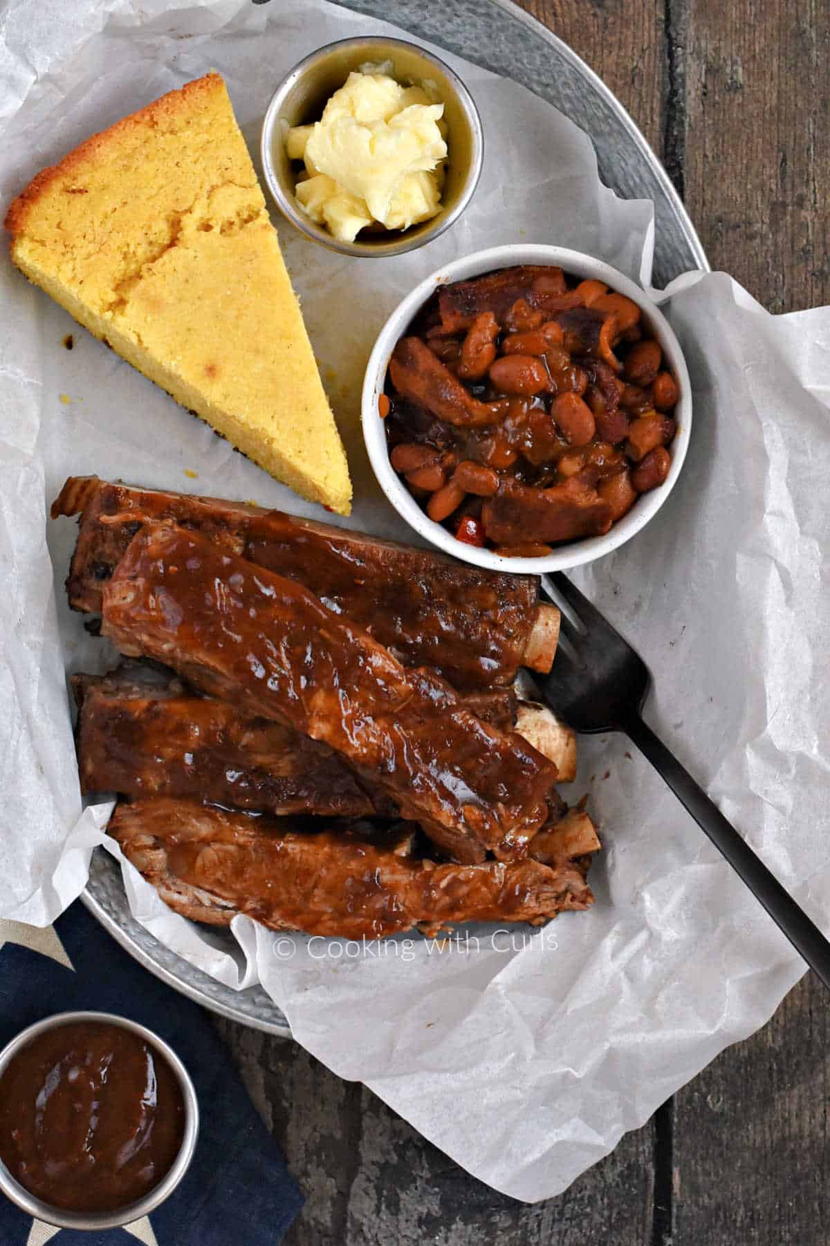 Looking down on a tray with barbecue ribs, cornbread wedge, and a small bowl of baked beans with bacon.