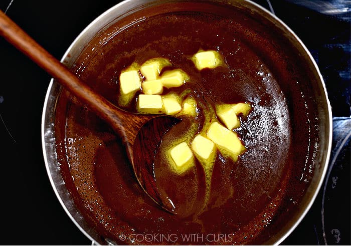 looking down on butter cubes melting into the hot chocolate gravy with a wooden spoon leaning against the left side of the saucepan. 