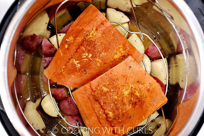 Looking down on two salmon filets on a wire rack over quartered red potatoes and chicken stock inside a pressure cooker. 