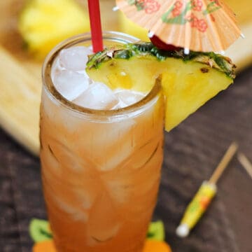 Orange cocktail in a tiki glass with a pineapple wedge, cherry, red straw and orange paper umbrella.