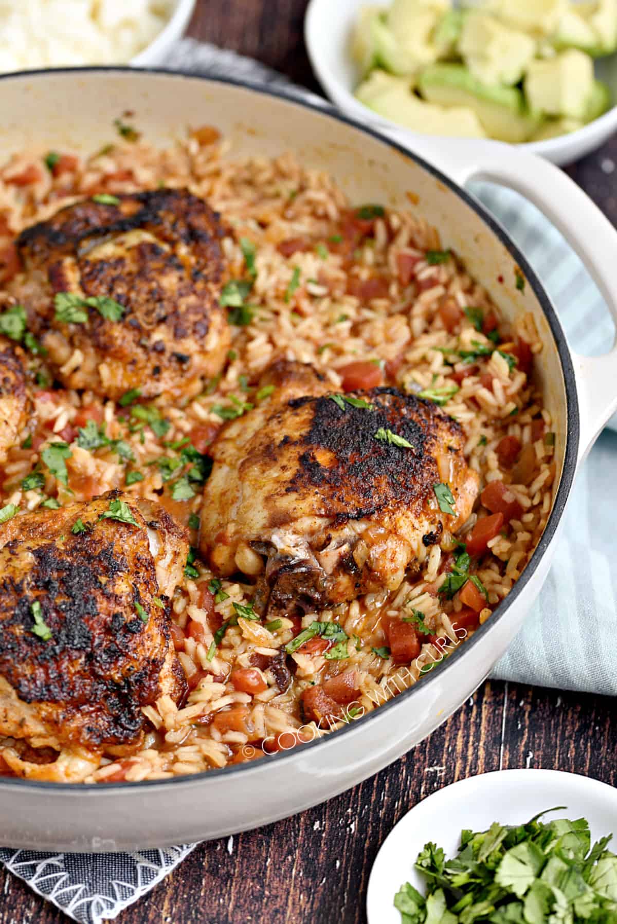 Chicken thighs cooked on a bed of Mexican rice in a white skillet with bowls of crumbled cheese and diced avocado in the background.