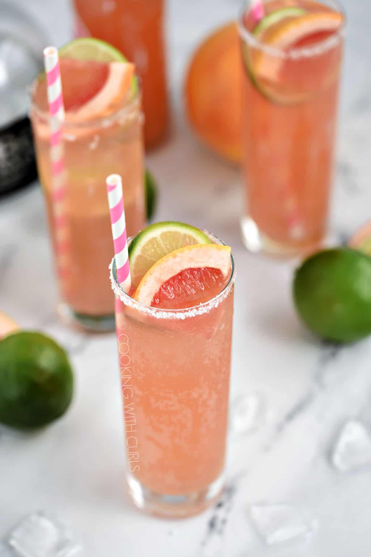 Three bubbly, pink cocktails in tall glasses garnished with a slice of lime and grapefruit with a pink and white striped straw.