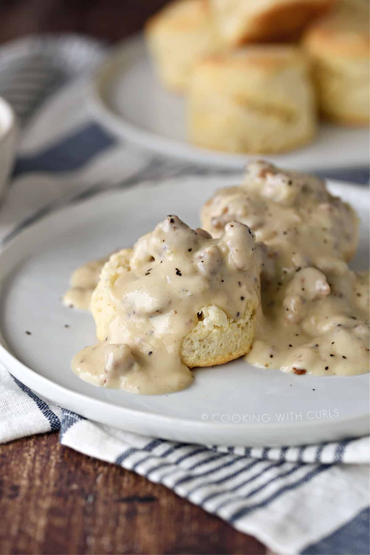 a biscuit split in half on a white plate topped with sausage gravy with a plate of biscuits in the background.