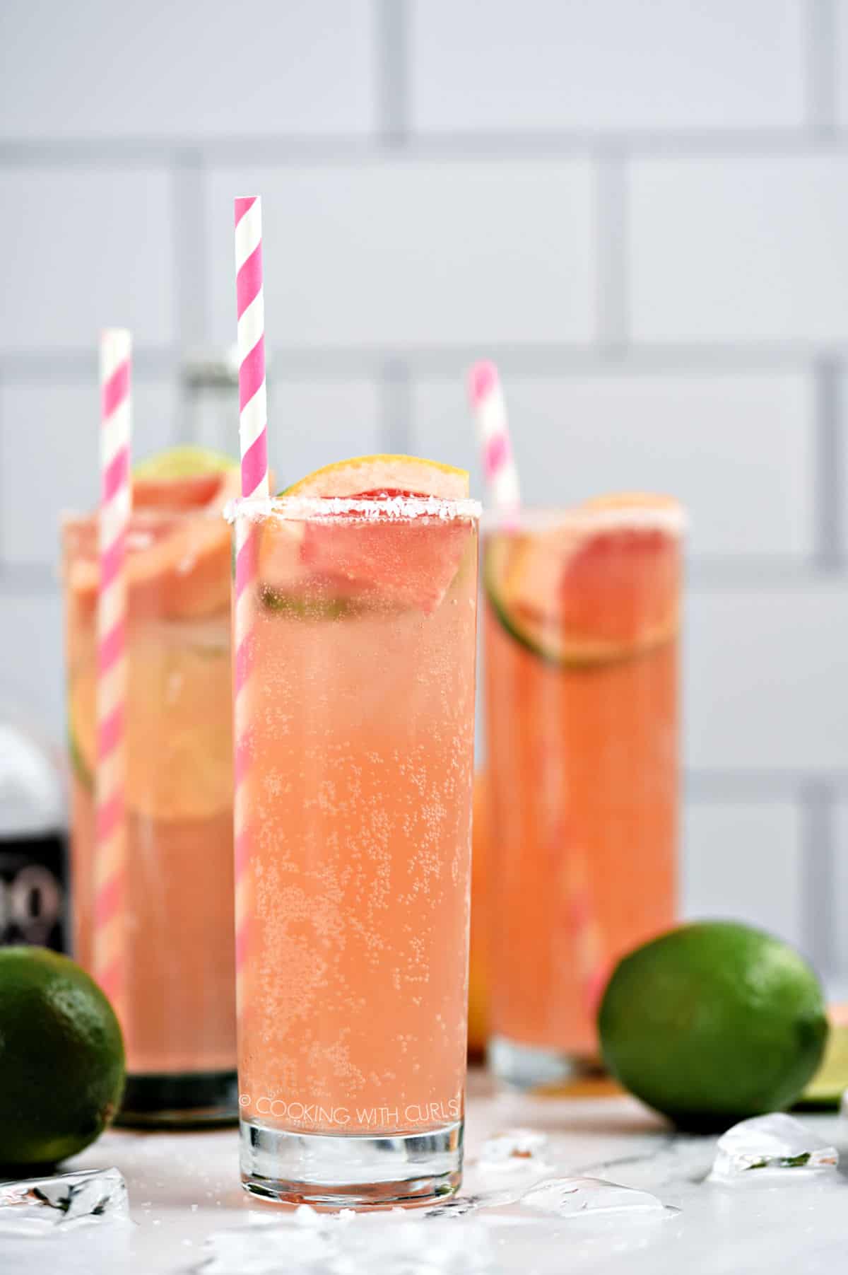Three bubbly, pink Paloma cocktails in tall glasses garnished with a slice of lime and grapefruit with a pink and white striped straw.
