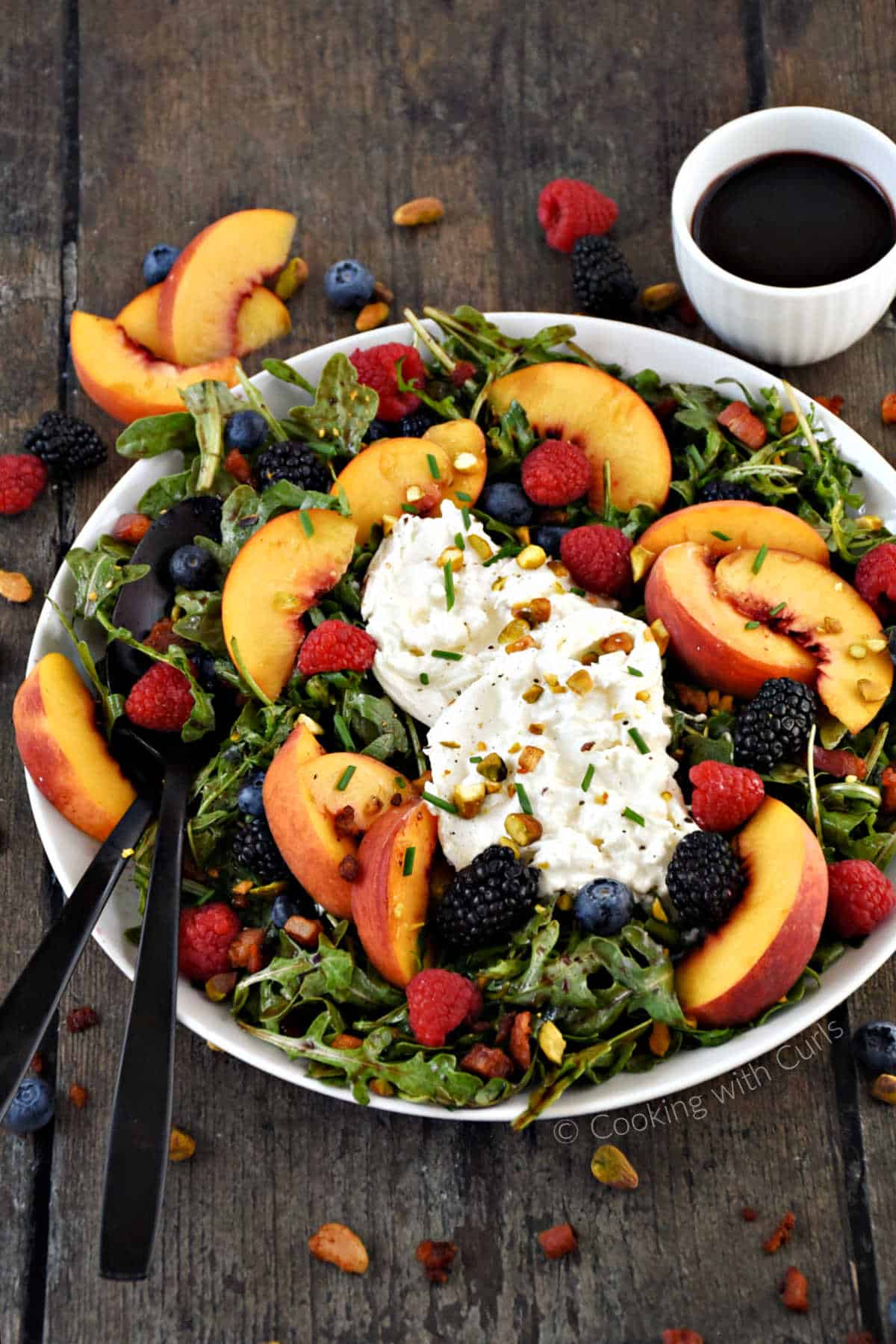 Sliced peaches, fresh berries, and fresh burrata on a bed of arugula with a bowl of vinaigrette on the side.