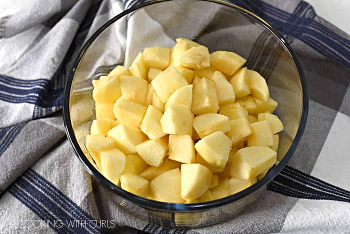 peeled, chopped apples in a glass bowl sitting on a blue, gray and white striped dish towel. 