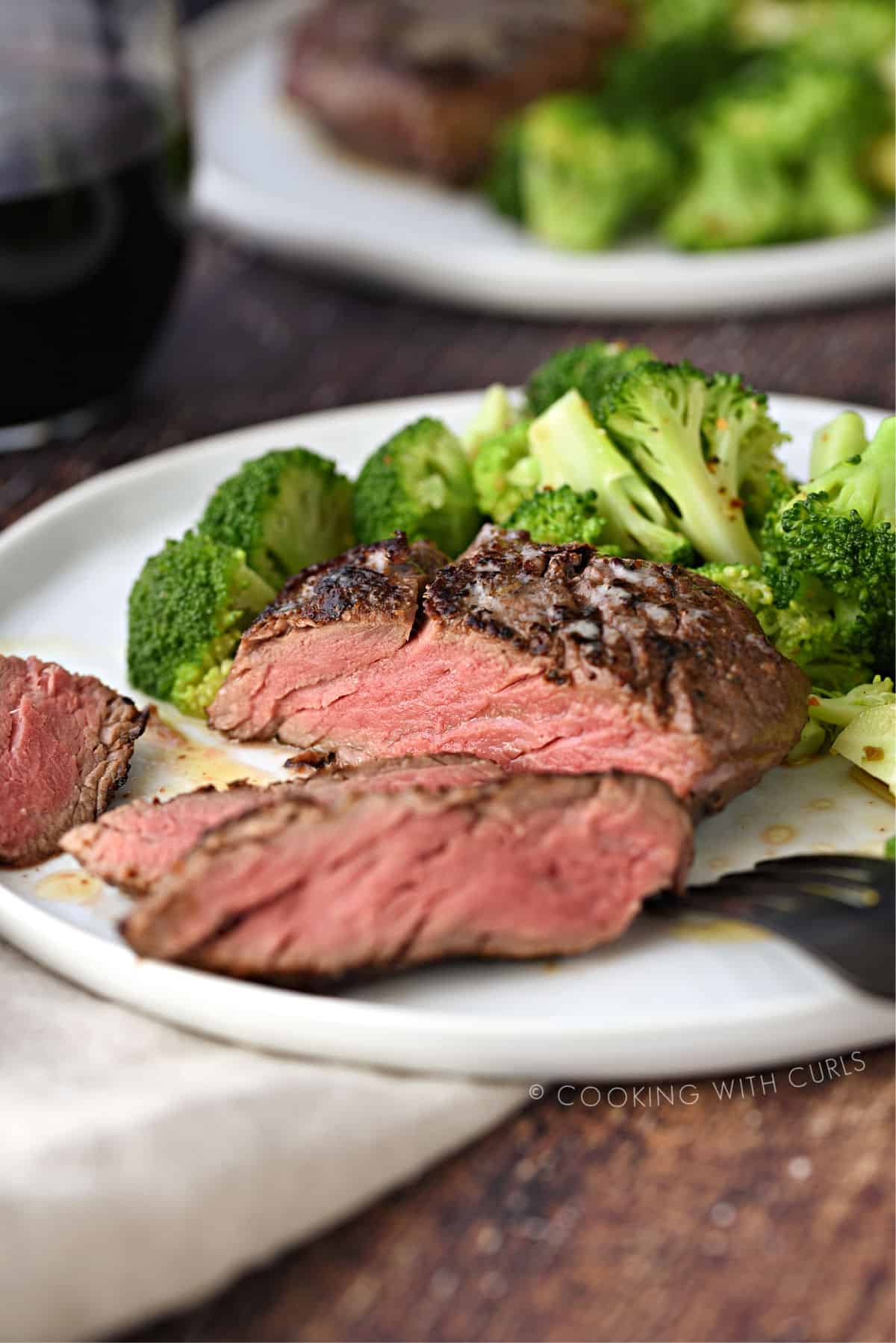 sliced filet mignon on a white plate with broccoli florets, with a second place and a glass of red wine in the background.
