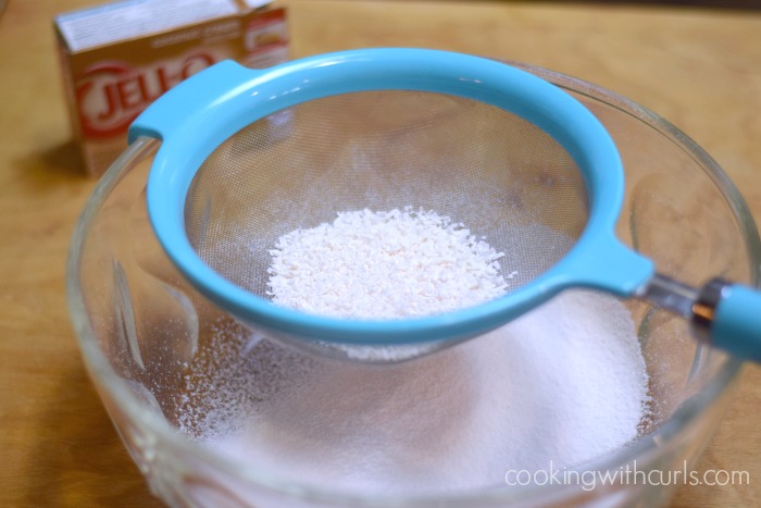 Coconut flakes in a blue sifter above coconut instant pudding mix in a large glass bowl.