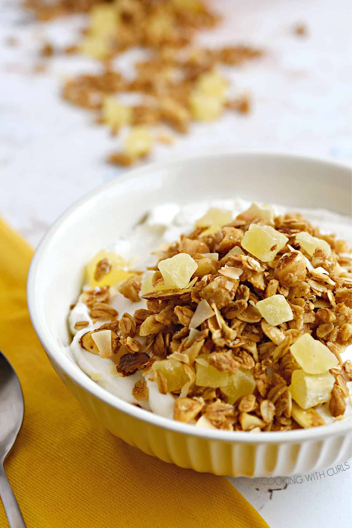 Pina Colada Granola topped yogurt in a white ribbed bowl sitting on a yellow napkin with a spoon to the left and spilled granola at the top.