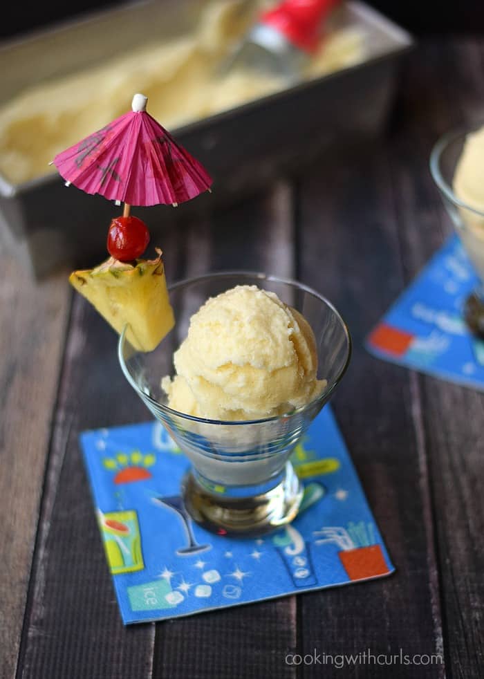 Piña Colada Ice Cream in a small glass garnished with a piece of pineapple, a cherry and pink paper umbrella.