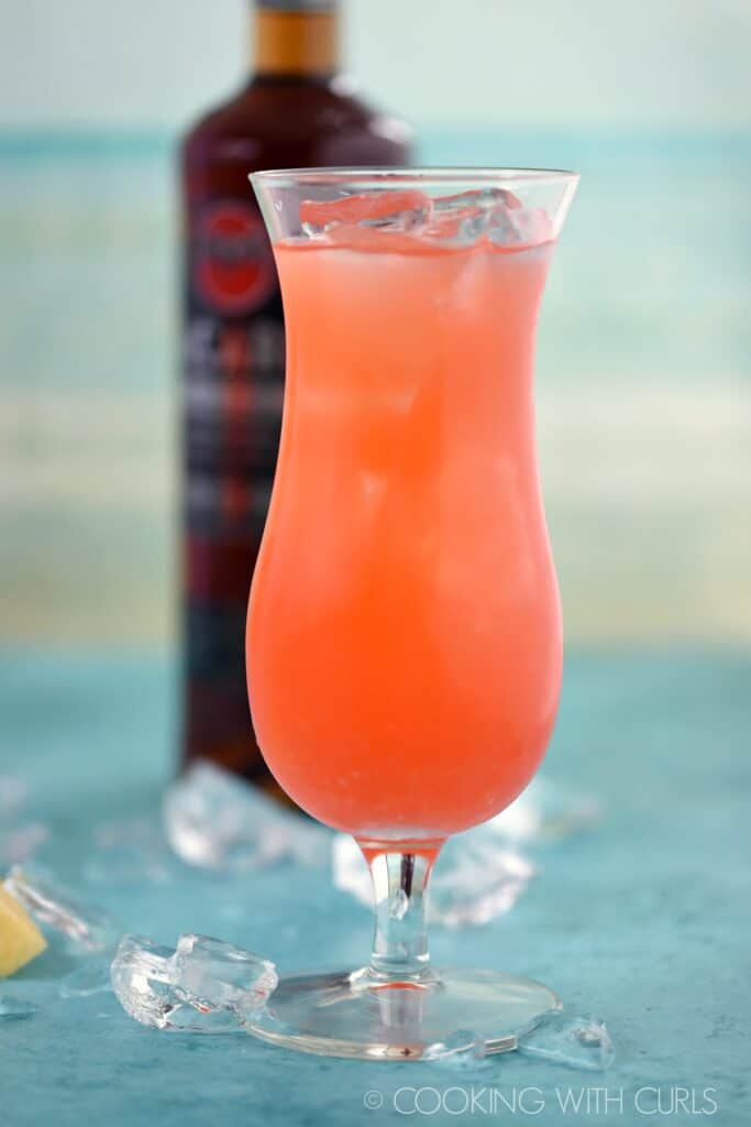 pink colored cocktail in a hurricane glass filled with ice with a bottle of dark rum in the background.