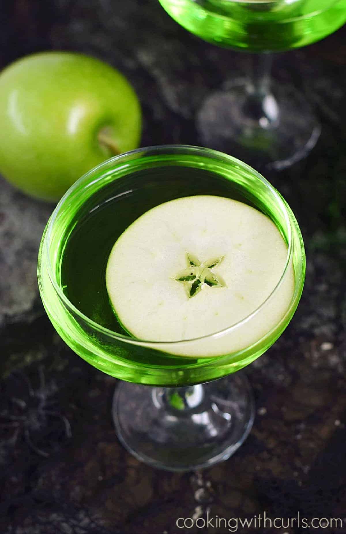 Looking down on a green cocktail with a floating apple slice with a second drink in the background with a green apple.