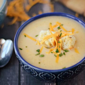 A dark blue bowl filled with potato cheese soup, and topped with croutons and shredded cheddar cheese with additional cheese in the background.