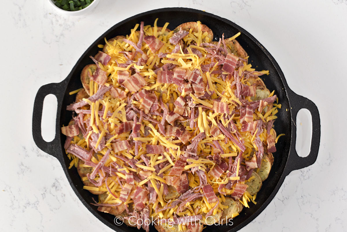 Potato slices, shredded cheddar cheese, corned beef and bacon pieces in a cast iron skillet. 