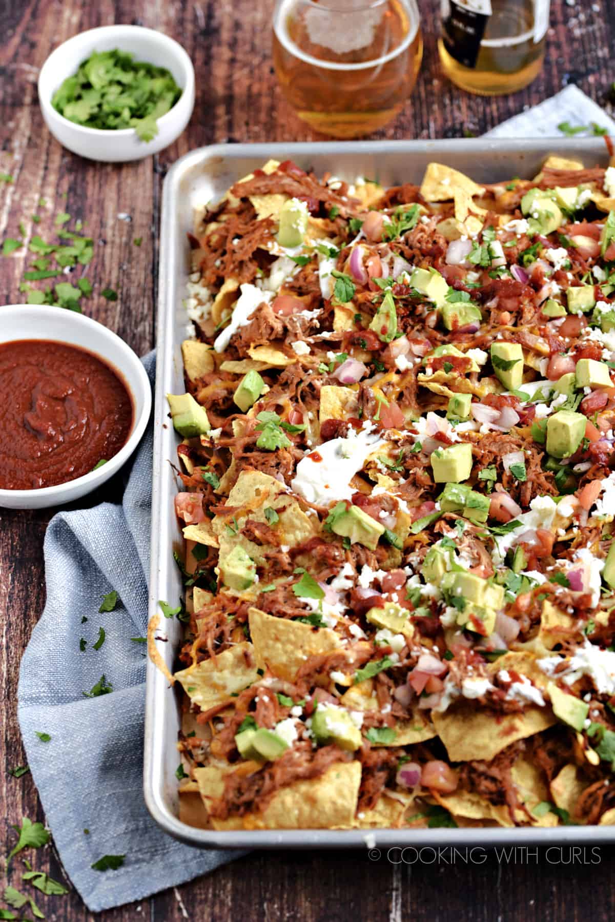 A sheet pan filled with Pulled Pork Nachos topped with avocado, sour cream, salsa and barbecue sauce. 