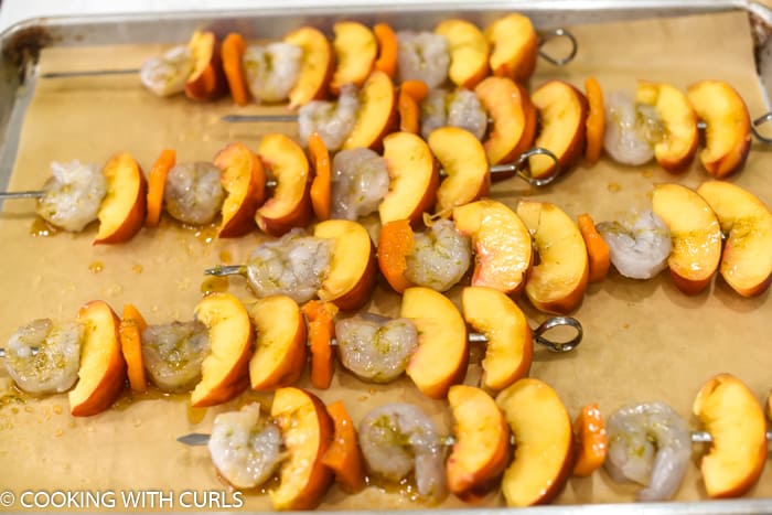 raw shrimp, bell pepper and peach slices on metal skewers laying on a parchment lined baking sheet. 