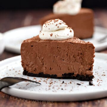 A slice of chocolate cheesecake topped with whipped cream and sprinkled with shaved chocolate sitting on a white plate with a second slice in the background.