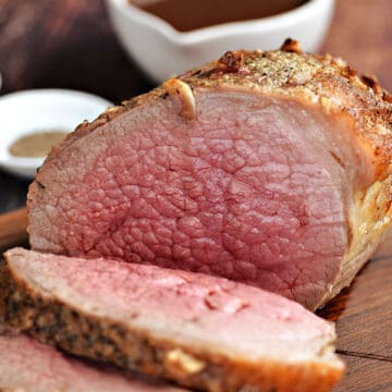 Medium-rare roast beef sliced on a dark wood cutting board with a bowl of gravy in the background.